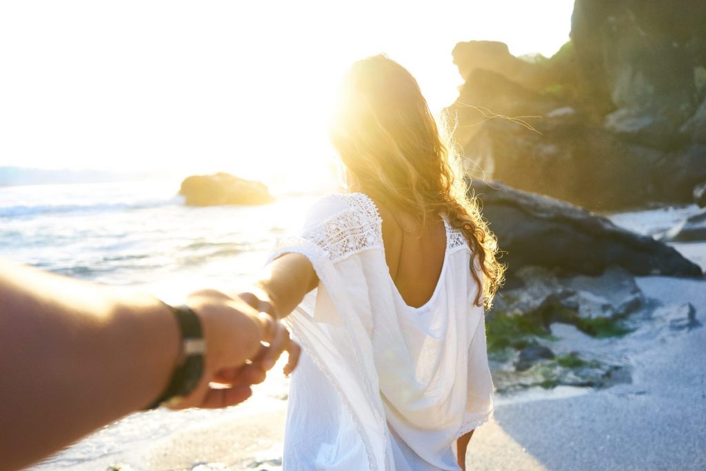 Person holding woman's hand beside sea while facing sunlight.