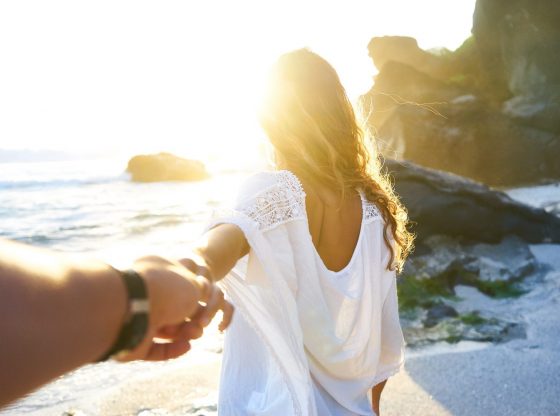Person holding woman's hand beside sea while facing sunlight.