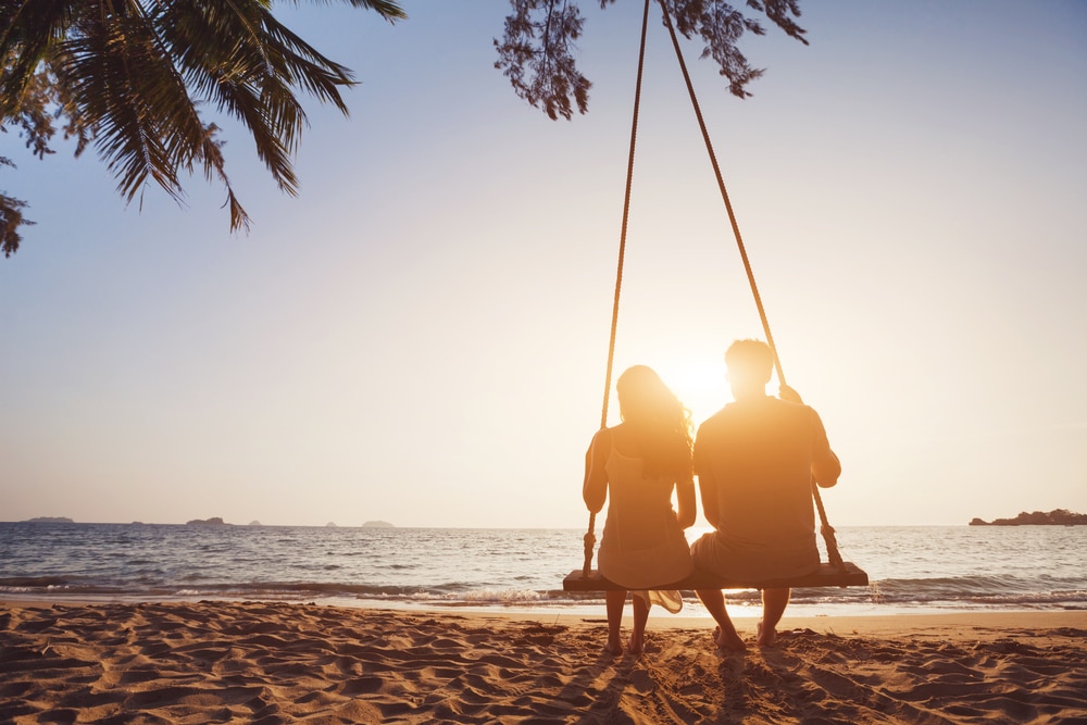 A couple on a swing on the beach watching the sunset.
