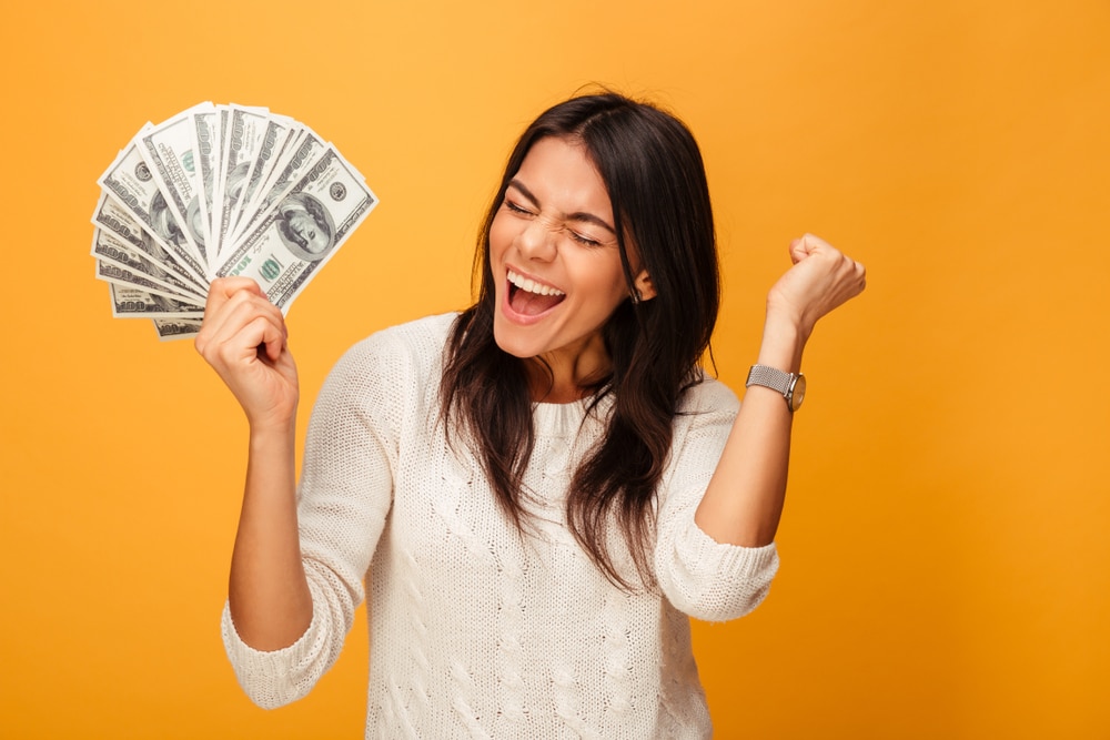 Portrait of a cheerful young woman holding and manifesting money and celebrating over yellow background