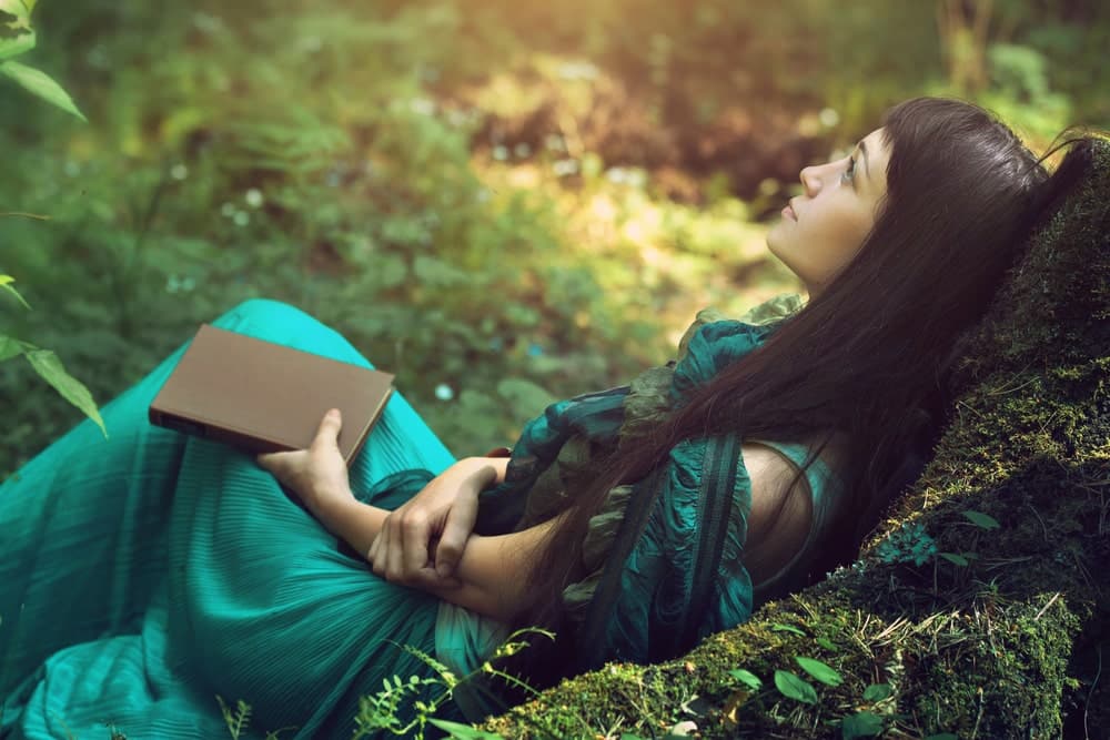 A Taurus woman with brown hair in a green dress sitting under a tree in the forest with a book