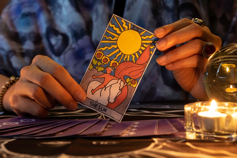 Close-up of a woman’s hands holding The Sun tarot card on a table near a burning candle and a crystal ball on World Tarot Day.