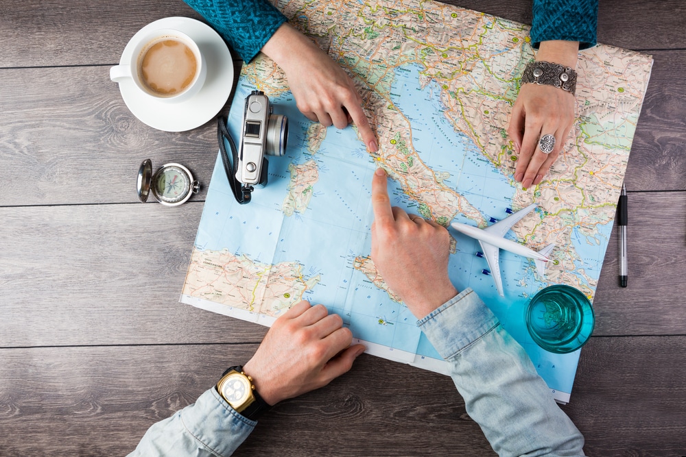 Top view of two pairs of hand pointing to a map with a coffee, pen, and compass on a table