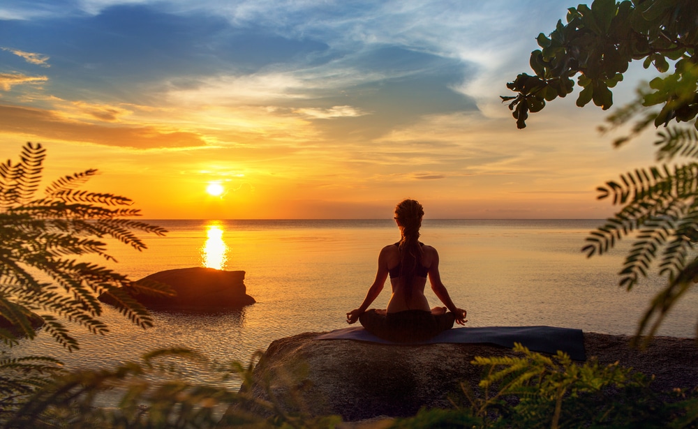 A woman meditating while looking at the ocean at sunset
